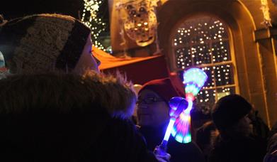A small boy, wearing a knitted hat and warm furry coat, is holding a multicoloured light up toy. He is in a crowd of people, who are standing in front
