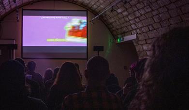 A purple-lit image of a groups of people facing a large screen, showing T-Blockers, a film directed by Alice Maio Mackay.