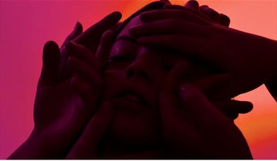 Close-up of a person's face surrounded by several hands, with dramatic red-pink lighting creating a moody, intense feel.