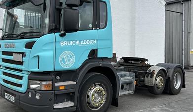A Picture of a large lorry front cab decorated in Bruichladdichs turcoise blue with a Bruichladdich whisky distillery logo on it. It sits in front of 