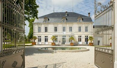 An image of the gated front to a 18th century french cognac chateau. The building is a large stone 3 floor chateau with a circular pool in front and a