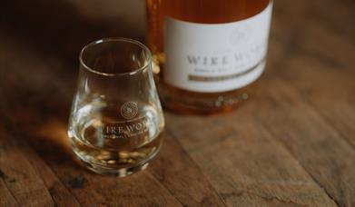 A small wire works branded glass containing single malt sitting on a wooden barrel top. In the background is a bottle of wire works single malt with a