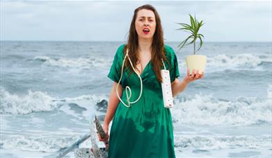 A woman looking perplexed is on a beach, she holds a ladder, a plant and a plug adapter.