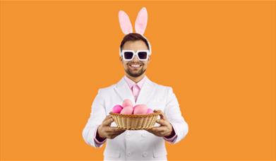 A cheerful man wearing a white suit, pink shirt, and bunny ears headband holds out a wicker basket filled with pink Easter eggs. He is also wearing ov