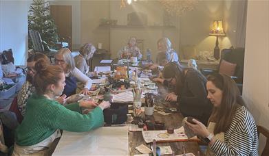 a seated group of people crafting at a large farmhouse table for the Love Makers Craft Cafe