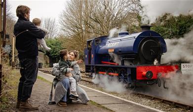 A family of four can be seen from behind waving at a blue steam train from the station platform.