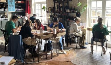 a seated group of people crafting at various tables in a spacious room lined with shelves of wine