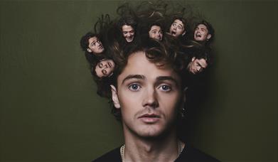 A photo of the comedian Henry Rowley. He is looking into the camera. In his hair there are 7 heads of Henry, they all have different facial expression