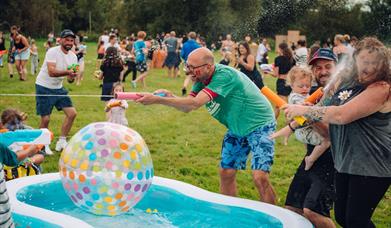 A group of people, including adults and children, are having a lively water fight outdoors on a grassy field.