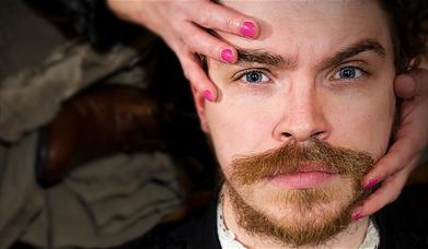 A white man with facial hair stares directly into the camera, while two hands with fingernails painted pink clasp his face.
