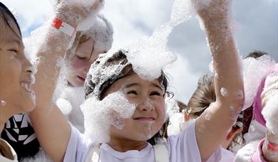 Children playing joyfully in a cloud of foam bubbles under a bright sky. A young girl in the center, smiling with arms raised, is covered in bubbles.
