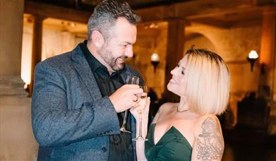 A smiling couple toast each other with a glass of Champagne around the Roman Baths