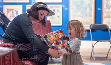 A lady reading wearing a headband with furry ears, reading a book to a young girl.