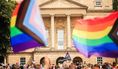 A photograph of the front of the Holburne Museum with a crowd of people in front. Two large progress rainbow flags fly in the foreground.
