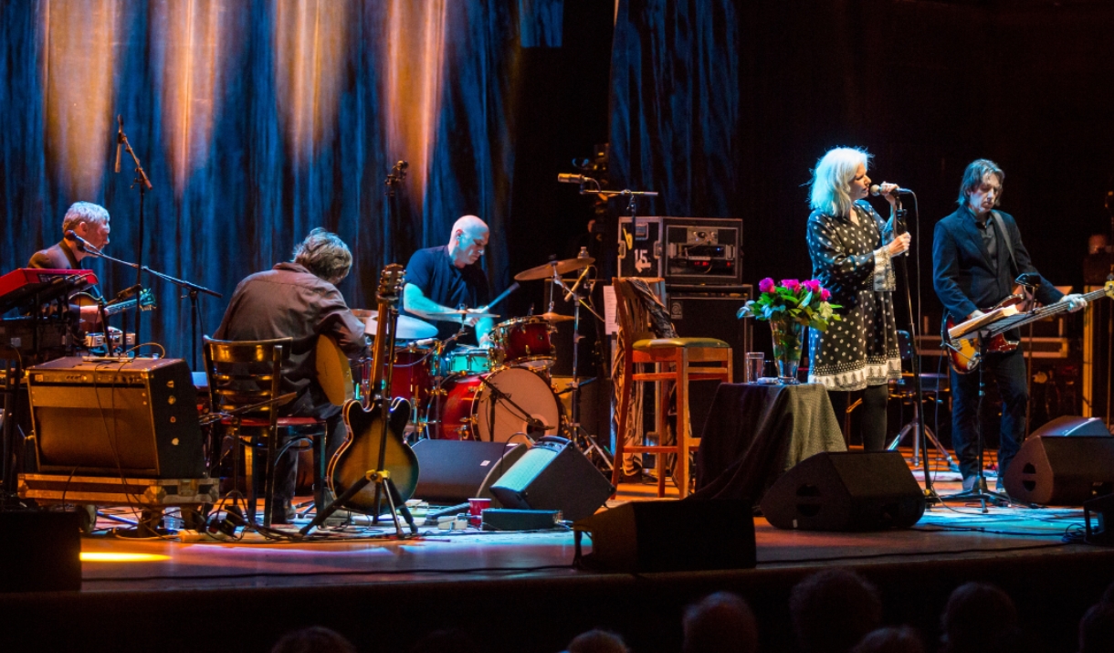 Band performing live on stage under warm lighting, with a female vocalist at the microphone and musicians playing guitar, keyboard, and drums.
