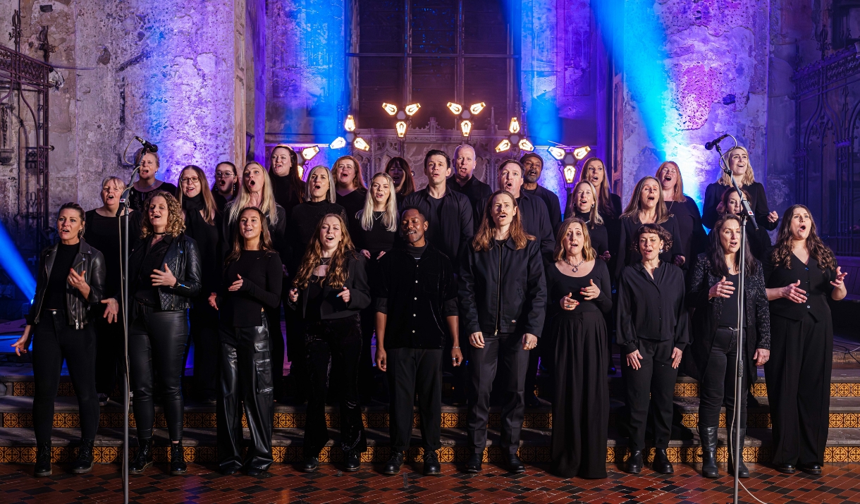 Large choir performing in a historic church, dressed in black and singing under blue and purple stage lighting.