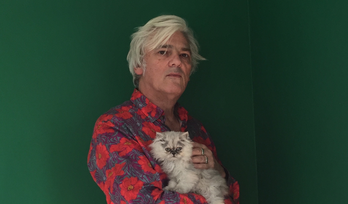 Robyn Hitchcock in a red floral shirt holding a fluffy kitten against a green background