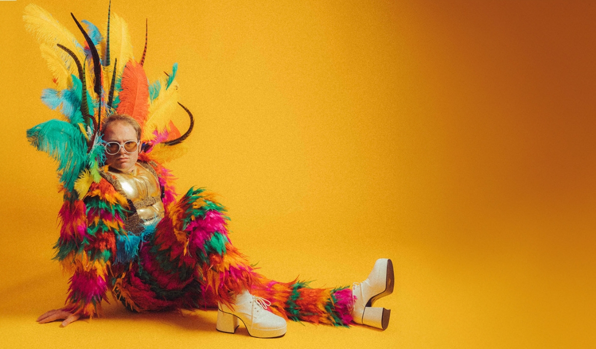 Person is a vibrant multicoloured feather costume posing on a yellow background.