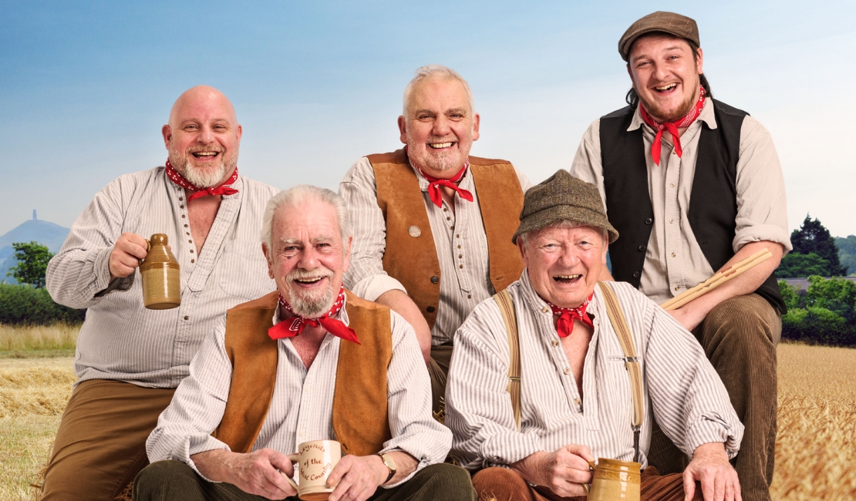 Six smiling men in traditional countryside clothing pose together in a wheat field, holding mugs, with hills and blue sky behind them.