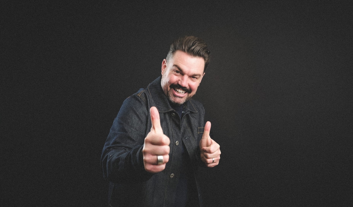 Portrait of Charlie Baker smiling in a dark jacket giving two thumbs up against a black background.