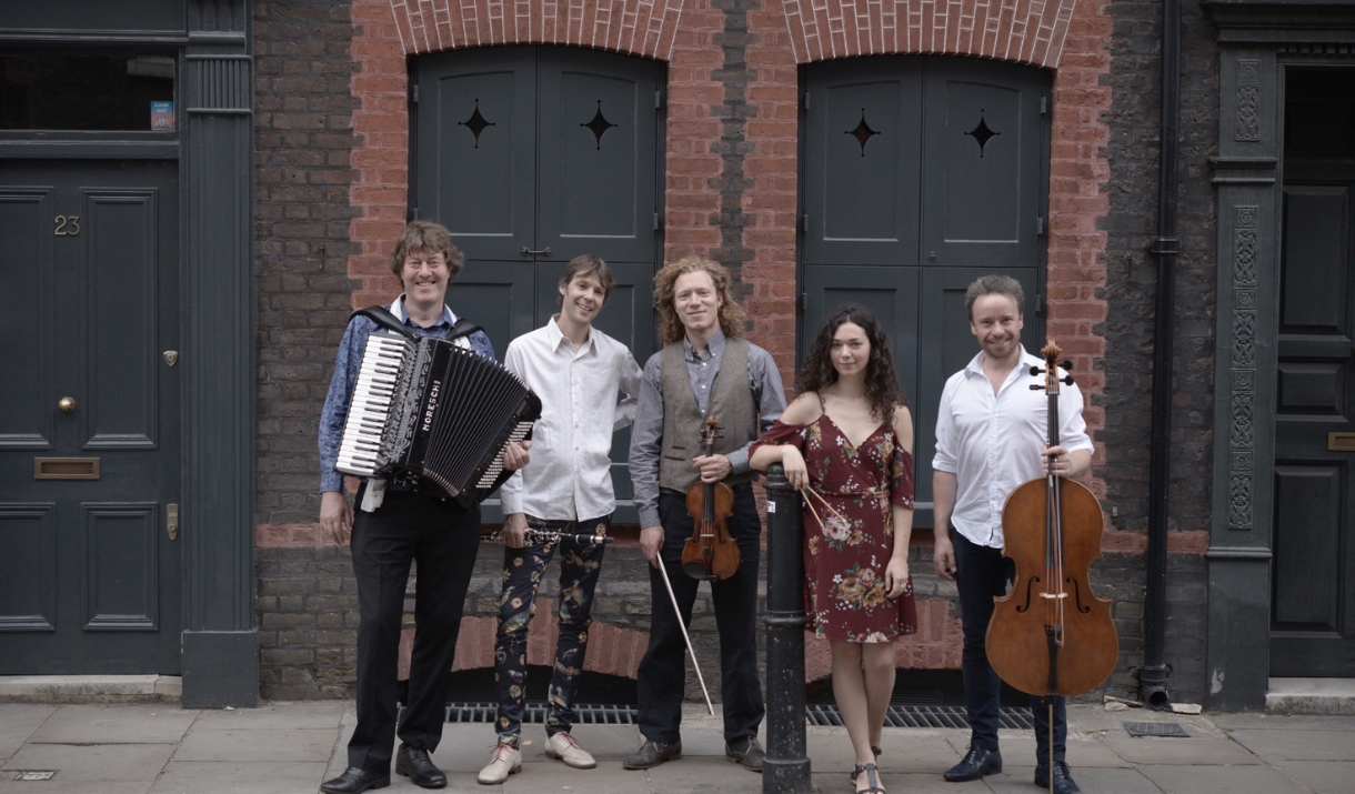 Five musicians standing outside brick buildings, holding accordion, clarinet, violin and cello, posing for a group photo.