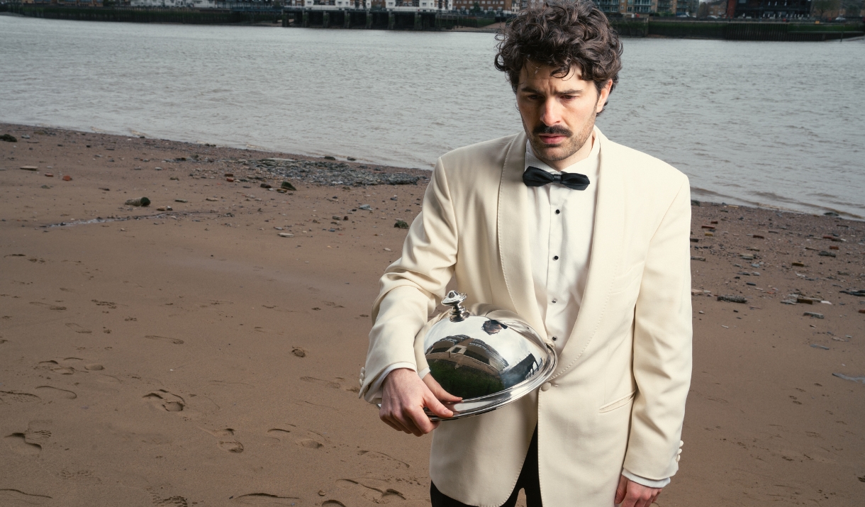 Paddy Young in a white jacket and bow ties holding a silver cloche on a sandy riverbank.
