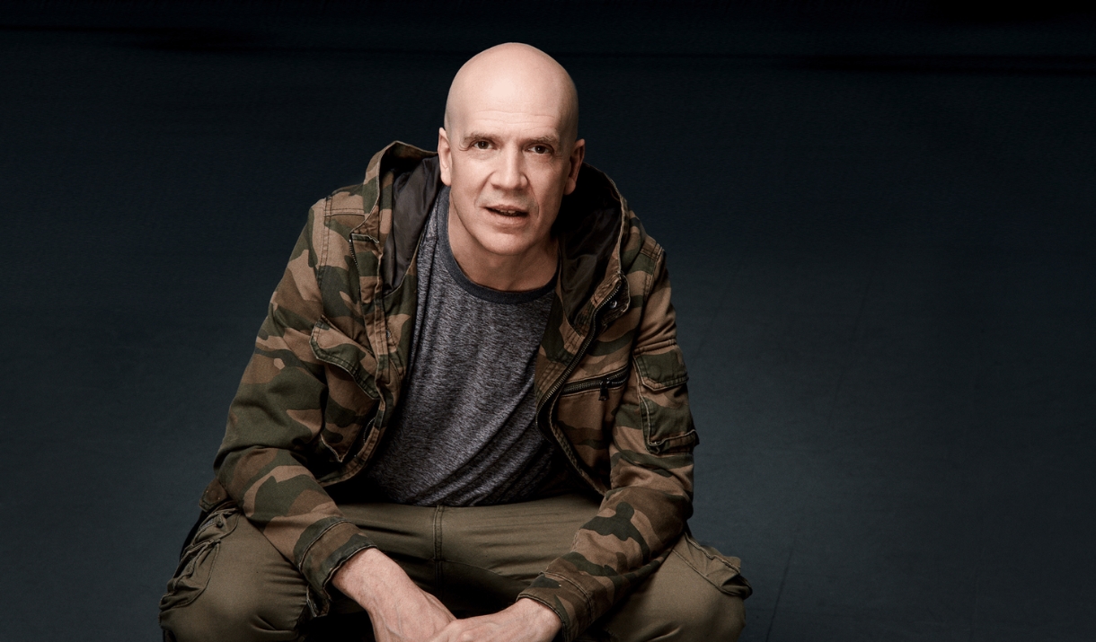 Devin Townsend in a camouflage jacket seated against a dark background.