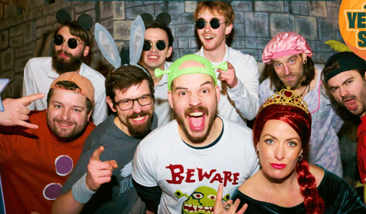 A group of people in Shrek-themed fancy dress, including Shrek, Princess Fiona, and various fairytale characters, grinning at the camera.