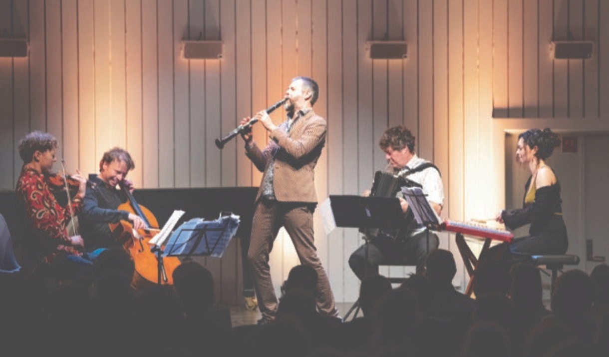 Live performance with five musicians on stage, including a clarinet, violin, cello, accordion and keyboard, playing to a seated audience.