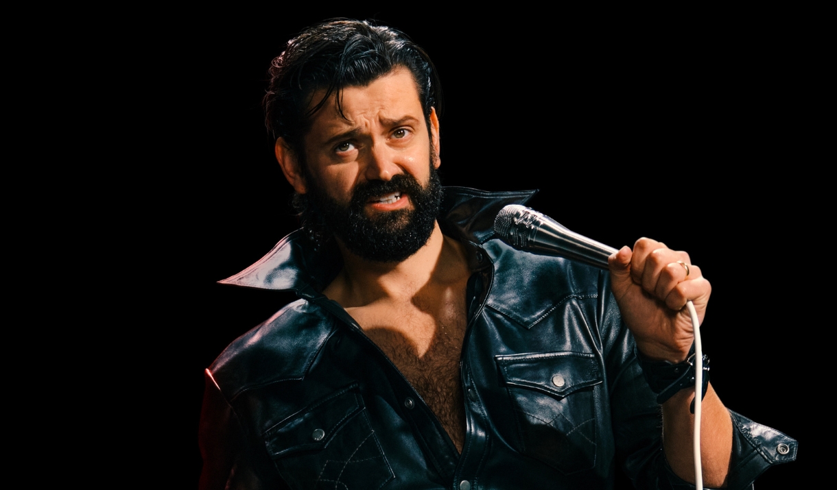 Fin Taylor in a black leather jacket holding a microphone against a dark background.