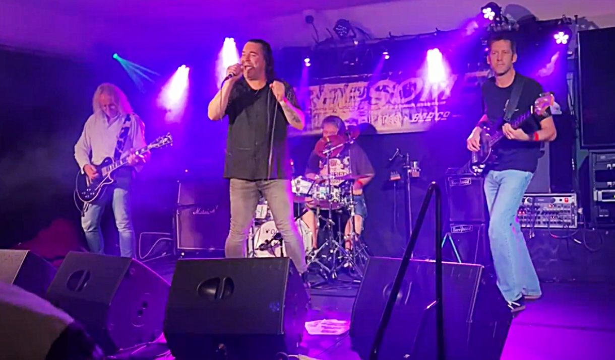 Live band performing on stage with singer, guitarist, bassist and drummer under purple lighting.