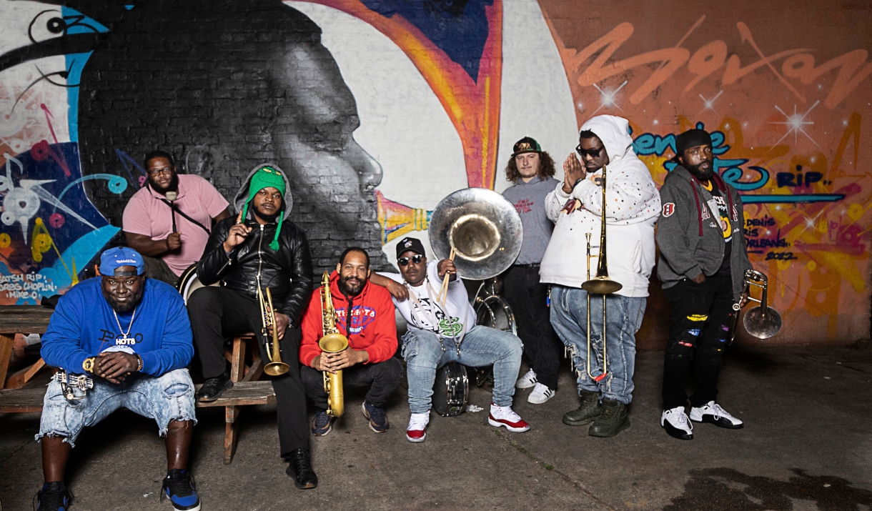Hot 8 Brass Band musicians holding saxophones, a sousaphone, and trombone, posed in front of a large graffiti mural at night.