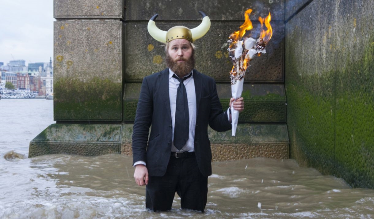 A man in a river wearing a smart outfit and a viking hat, holds a large piece of burning paper.