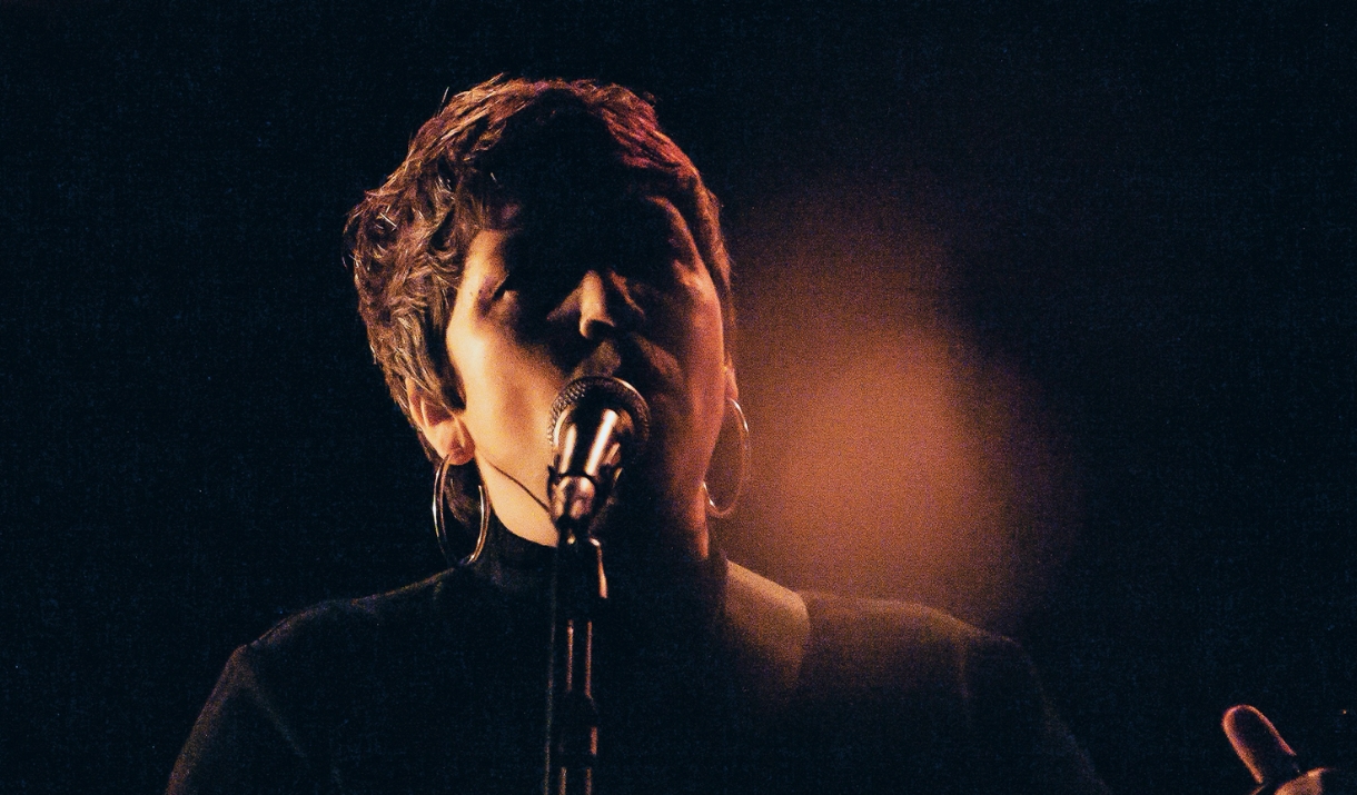 Singer performing into a microphone under warm stage lighting, close-up portrait.