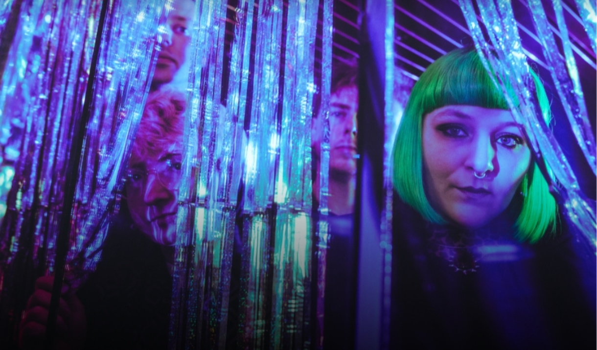 Four people peering through glittering tinsel curtains in purple and teal light.