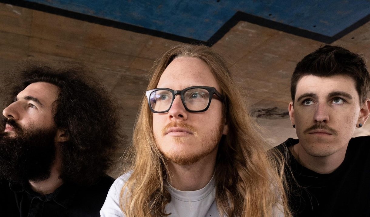 3 Men from the band Fantastic Stranger sit in front of a brick wall.