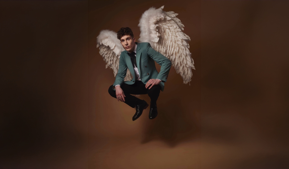 Larry in a green suit crouching mid-air with large white angel wings against a brown backdrop.