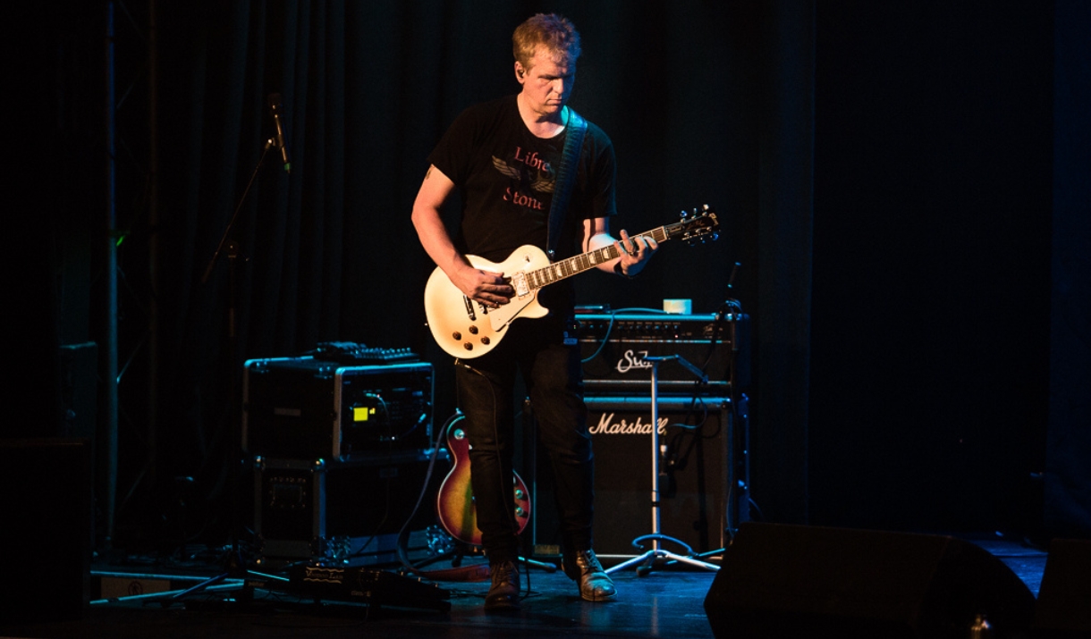 Dion Smith playing a cream Les Paul electric guitar on a darkened stage, with Marshall amps behind him.