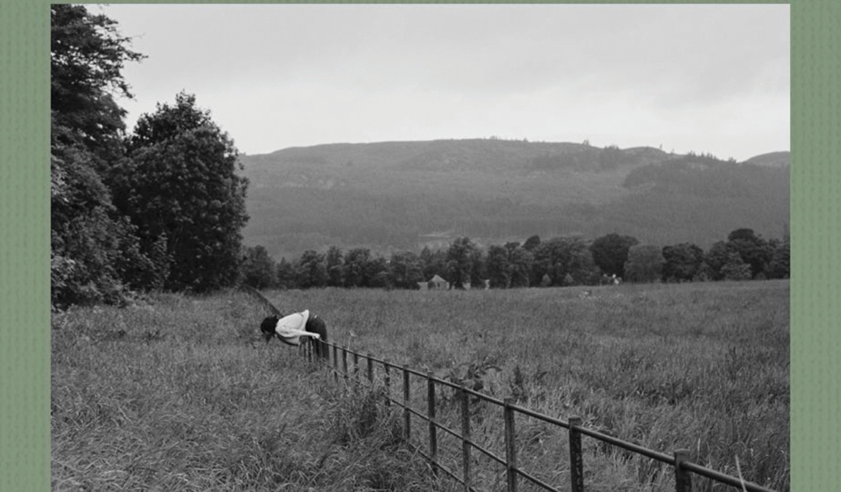 Black and White photograph of a person leaning over a rural fence in an open field, with trees and rolling hills in the background