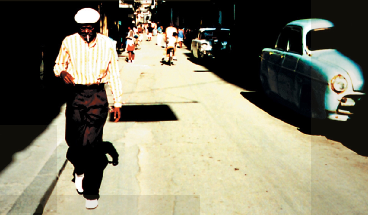 Man in striped shirt and cap walks down a sunlit street with vintage cars and pedestrians in the background.