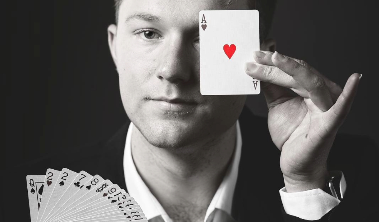 A black and white photo of the Magician Angus Baskerville, He is holding a deck of cards and covering one eye with the ace of hearts.
