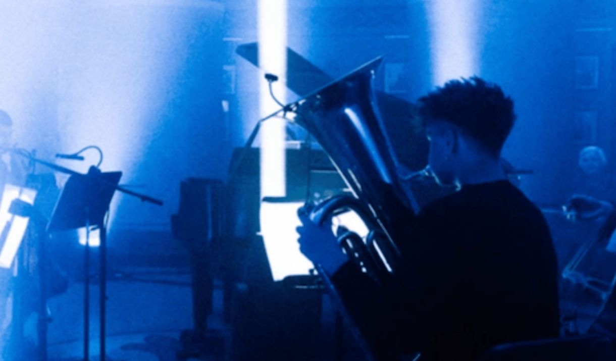 In a grandiose venue with Bath's iconic roman pillars, a  band is playing, focus on the brass player. Sparkfest logo in the top corner