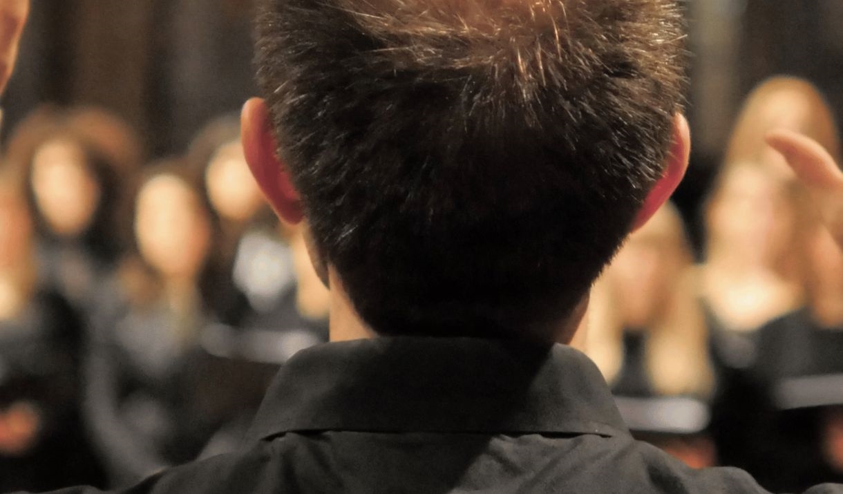 A composer stands with his back to the camera conducting a choir. The Sparkfest logo is in the top corner