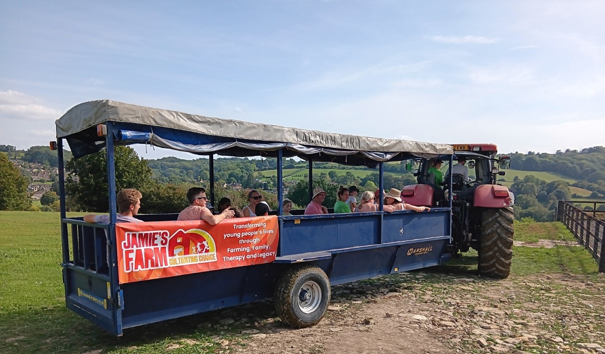 people sitting in a tractor trailer, in a green field, on a sunny day with an orange Jamie's Farm banner on the side of the trailer