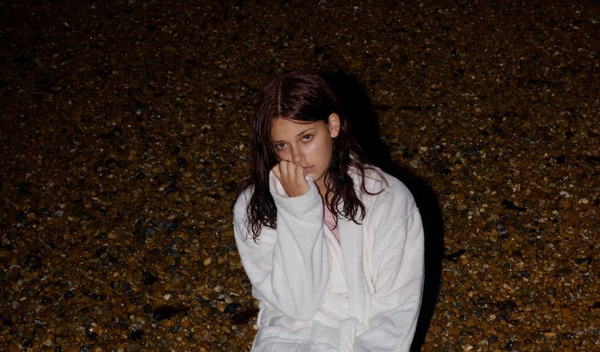 It's evening, a young woman in a white dressing gown sits down on a beach.