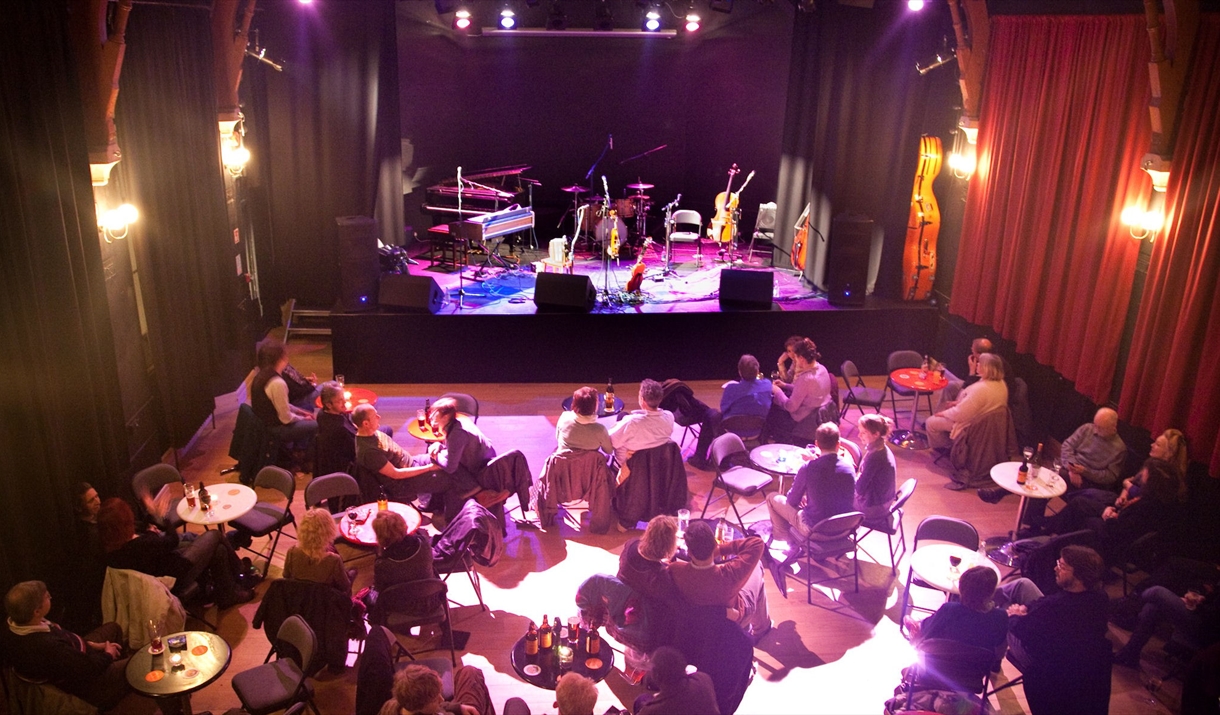 People sitting at individual tables in front of the stage at the Chapel Arts Centre in Bath