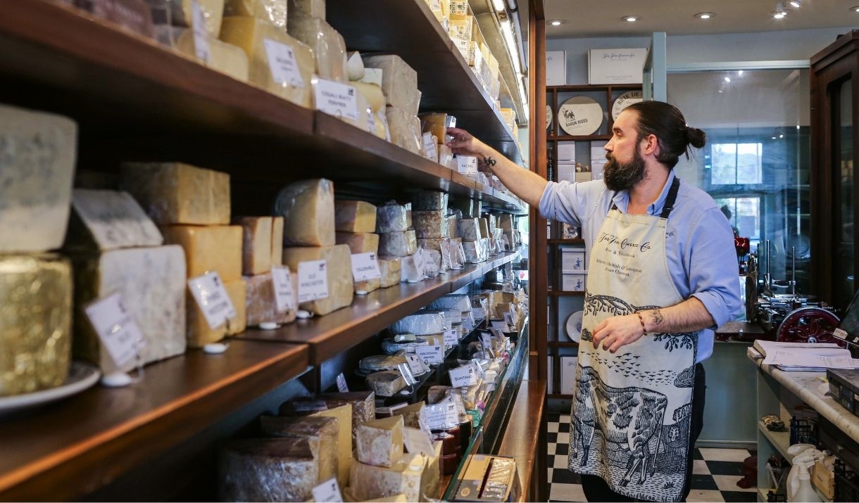 An employee wearing an apron decorated with a cow reaches towards a selection of cheeses on a shelf.