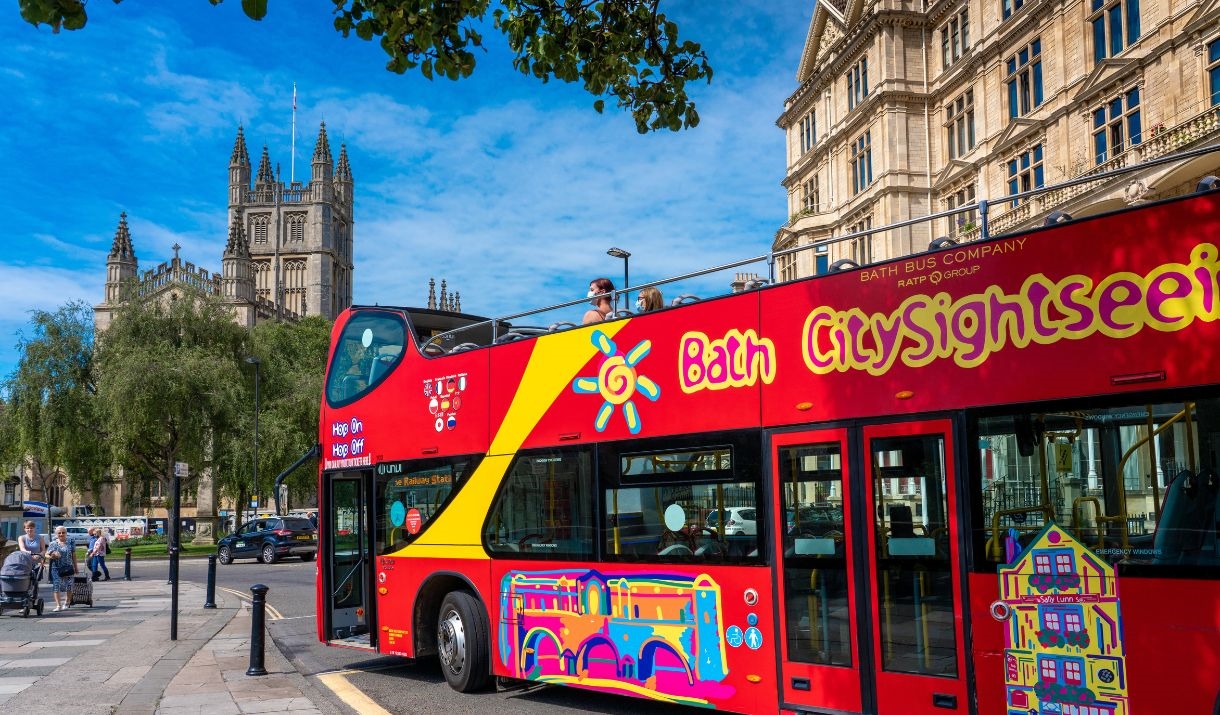 Red "Bath CitySightseeing" open-top bus with Bath Abbey visible in the background.
