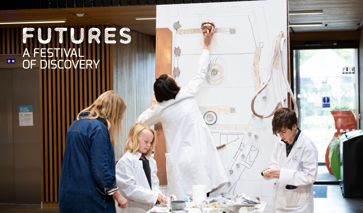A group of children wearing lab coats, along with a researcher facilitator are crafting a display of urban planning using household items.