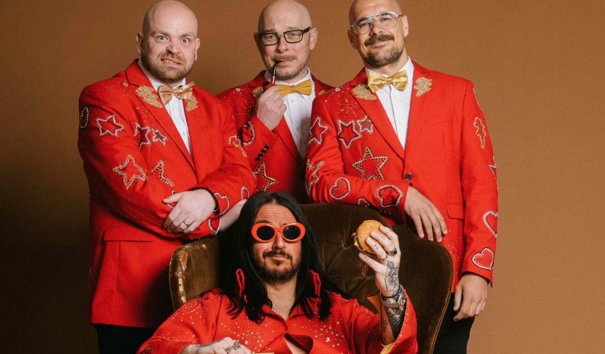 A photo of the band Elvana, they are clad in red, one of the singers in sitting on a chair holding a burger.